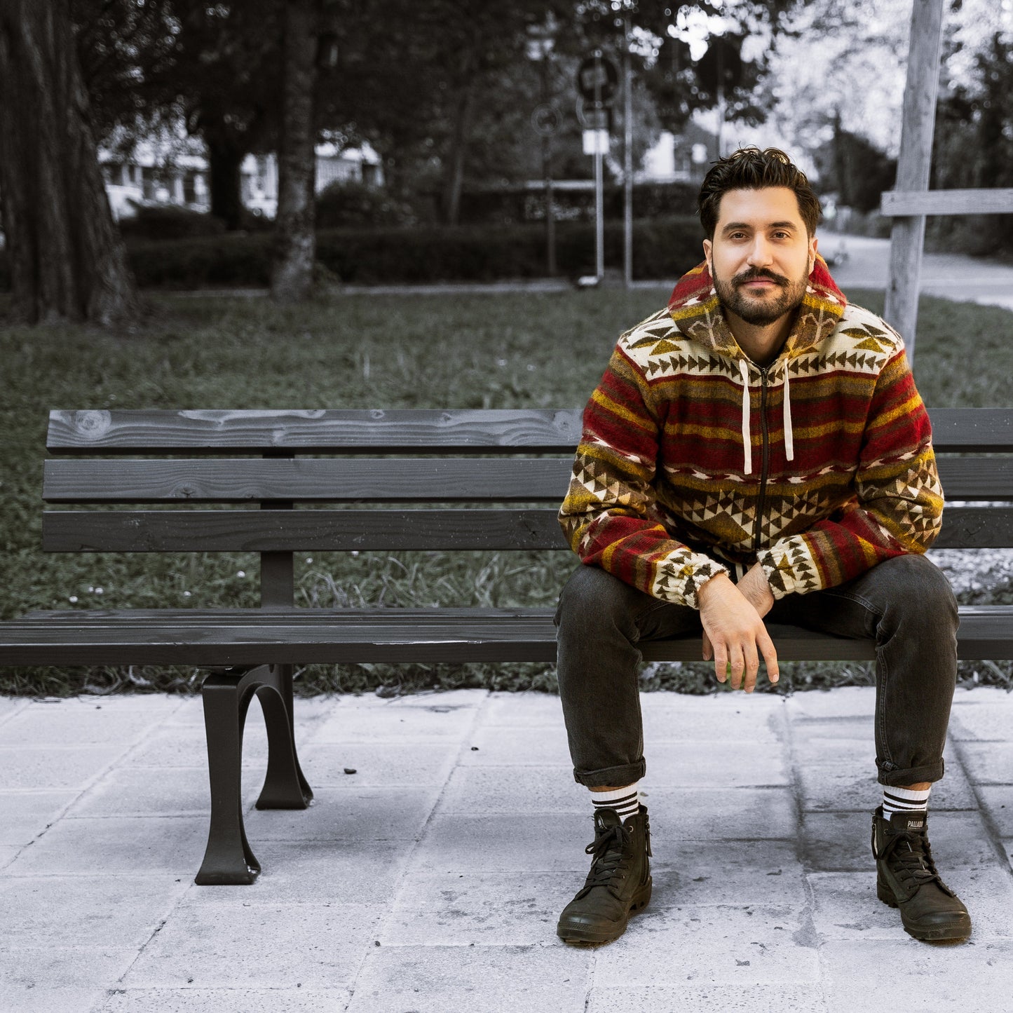 Front view of a man sitting on a bench wearing the ALLPA boho jacket made from a wool blend with alpaca, featuring red, brown, yellow, and cream geometric Andean patterns with a hood. Photographed outdoors in a park setting.