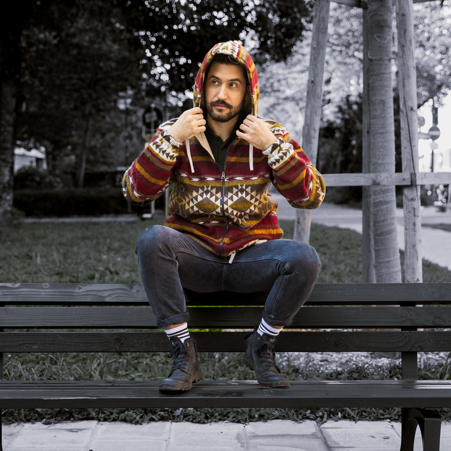Front view of a man sitting on a bench wearing the ALLPA boho jacket with the hood up. The jacket is made from a wool blend with alpaca and features red, brown, yellow, and cream geometric Andean patterns. Photographed outdoors in a park setting.