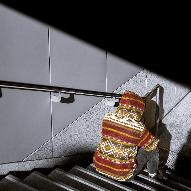 Back view of a man wearing the ALLPA boho jacket with hood up, made from a wool blend with alpaca, featuring red, brown, yellow, and cream geometric Andean patterns. Photographed from above while sitting on stairs in an urban setting with dramatic lighting.