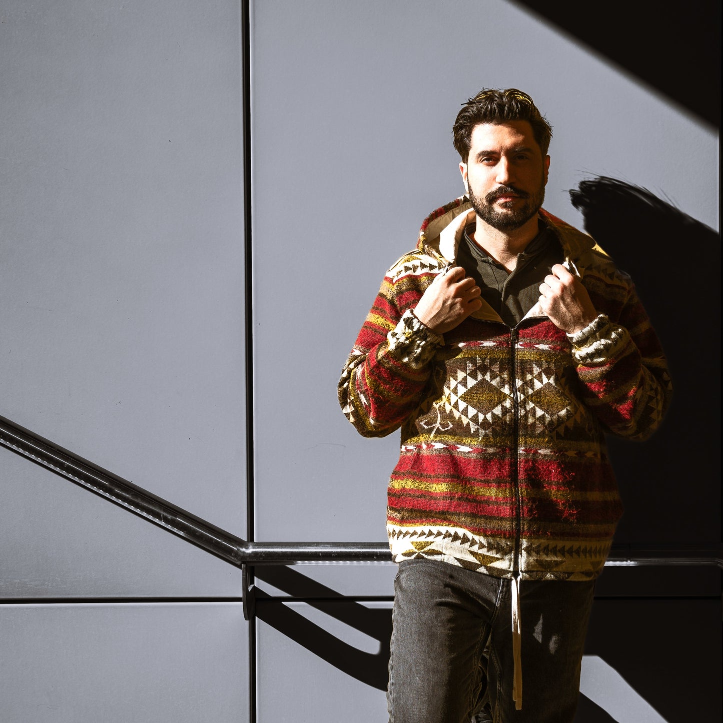 Front view of a man wearing the ALLPA boho jacket made from a wool blend with alpaca, featuring red, brown, yellow, and cream geometric Andean patterns. Photographed in an urban setting with dramatic sunlight and shadow contrast.