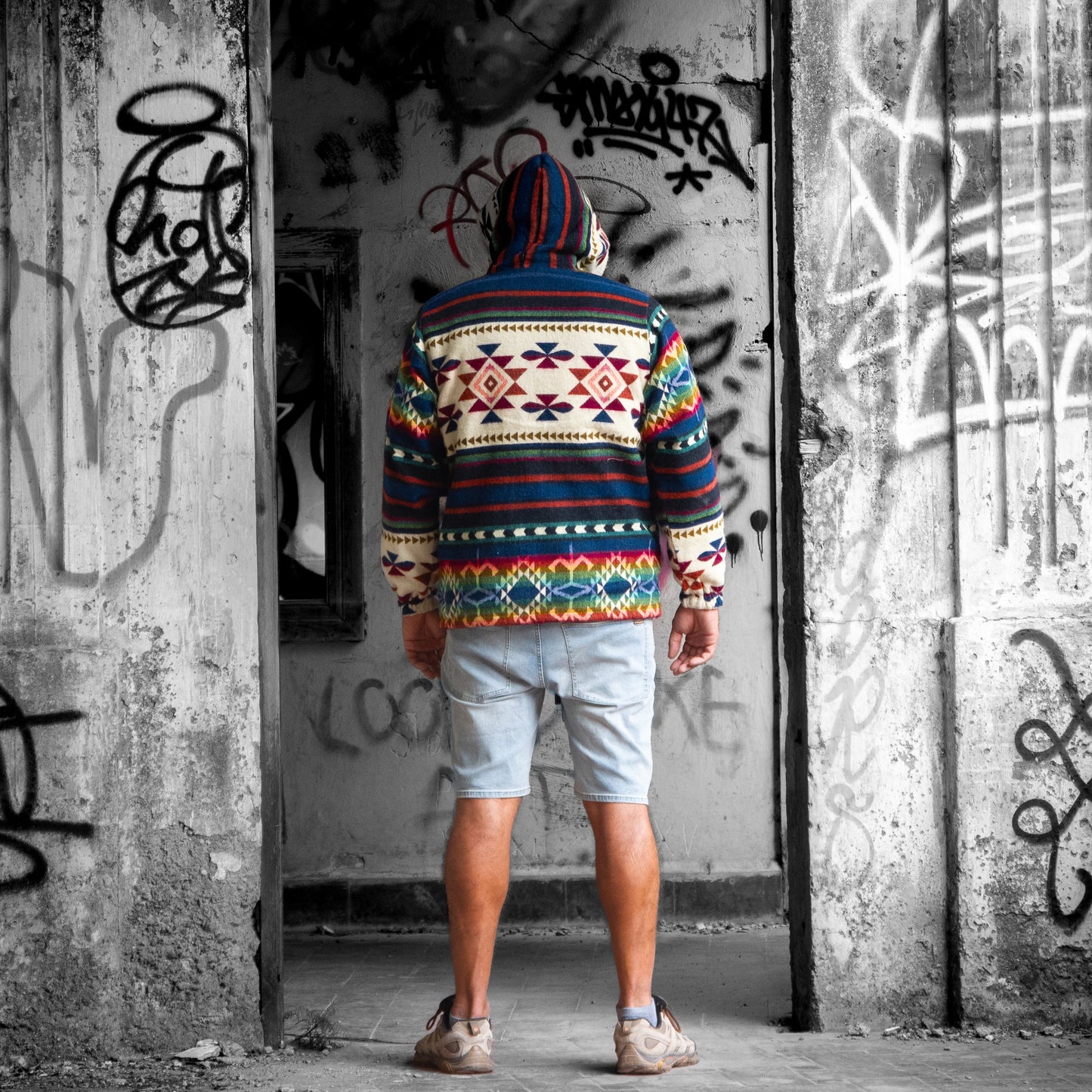 Back view of a man wearing the ANKA boho jacket made from wool blend with alpaca, showing colorful geometric Andean patterns and a hoodie. The model stands in front of a graffiti-covered wall in an urban setting.