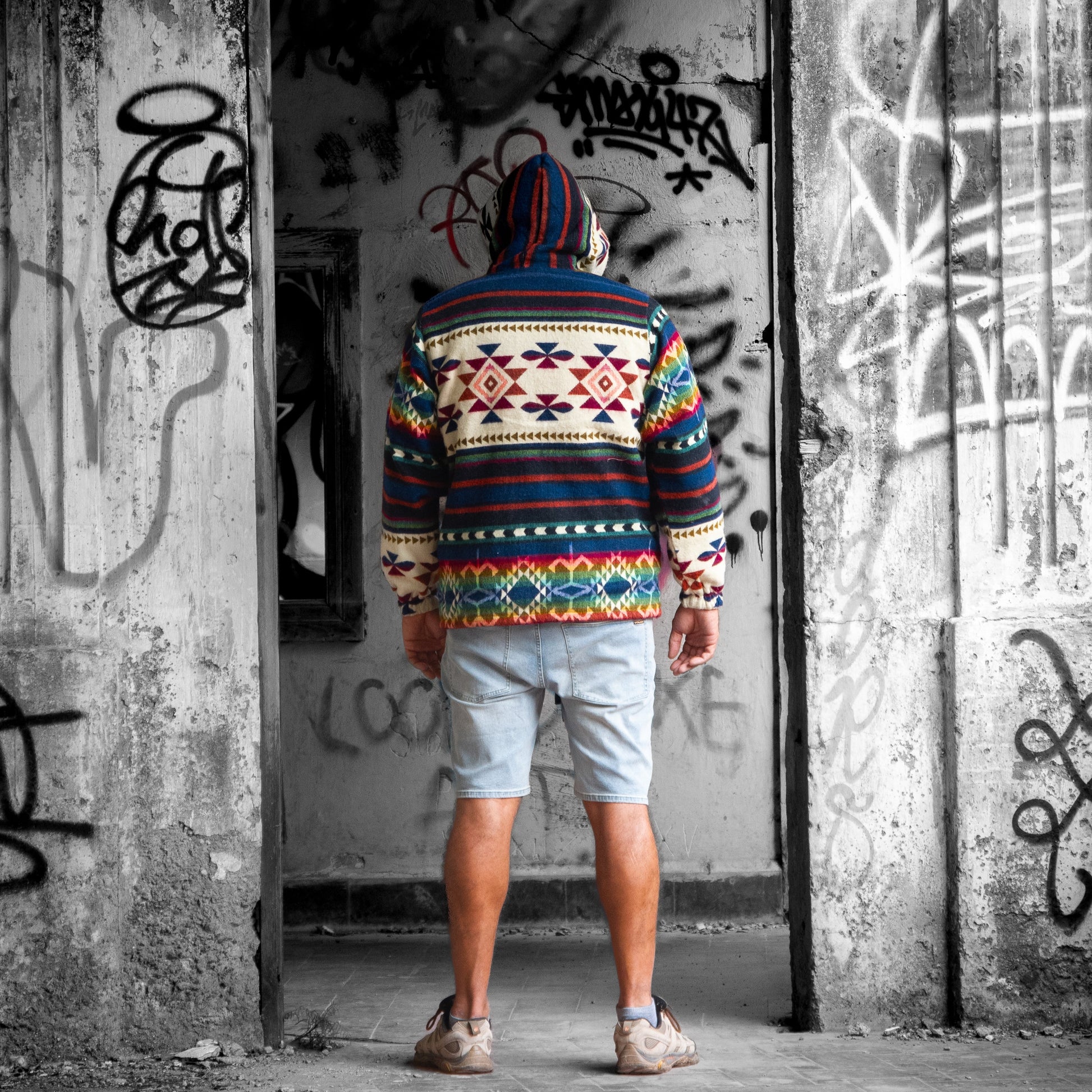 Back view of a man wearing the ANKA boho jacket made from wool blend with alpaca, showing colorful geometric Andean patterns and a hoodie. The model stands in front of a graffiti-covered wall in an urban setting.