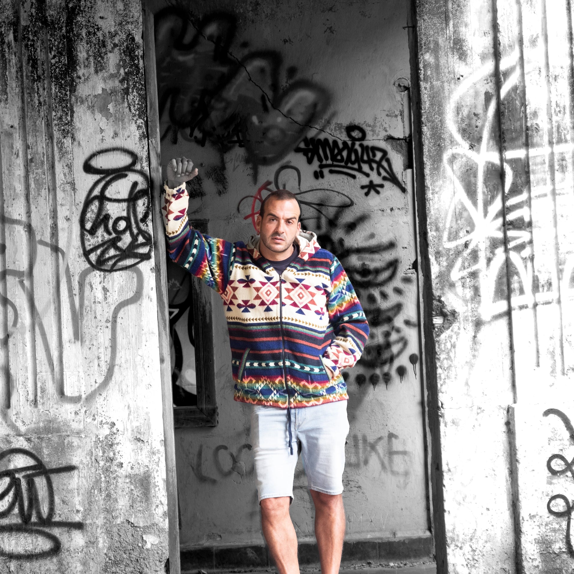 Front view of a man wearing the ANKA boho jacket made from wool blend with alpaca, posing against a wall with graffiti. The colorful geometric Andean patterns stand out in contrast to the black-and-white background.