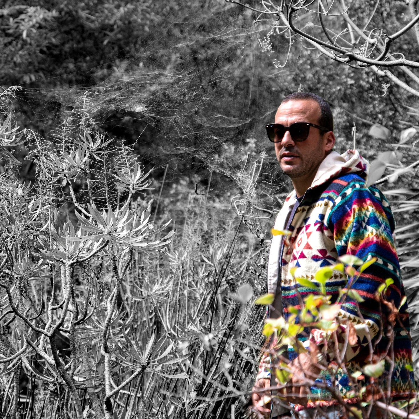 Man wearing the colorful Wayra boho jacket made of wool blend and alpaca, photographed outdoors among plants, featuring geometric Andean patterns and a hoodie with cream lining.