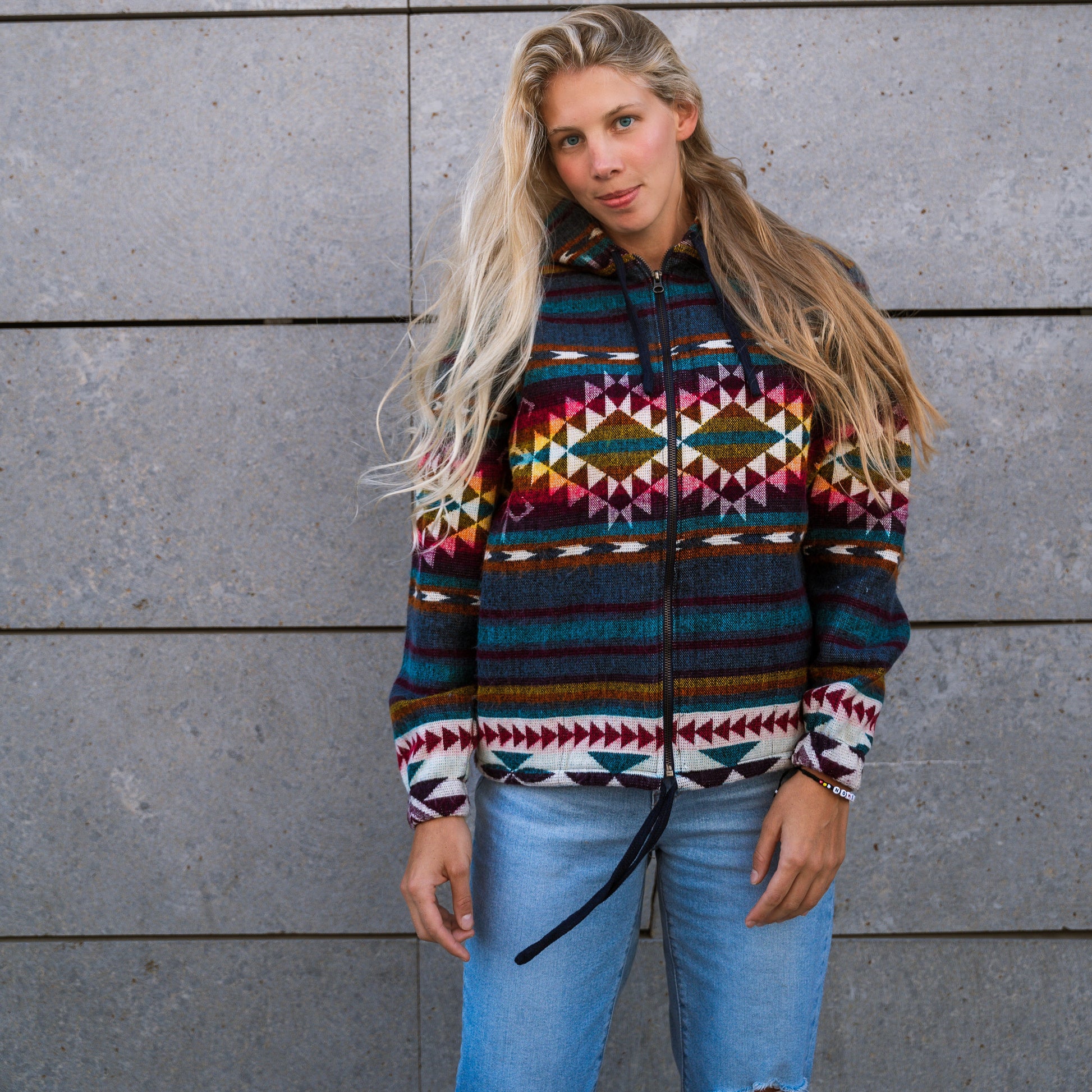 Front view of a woman wearing the QILLU boho jacket made from a wool blend with alpaca, featuring vibrant Andean geometric patterns in red, yellow, green, and cream. Photographed outdoors against a stone wall.