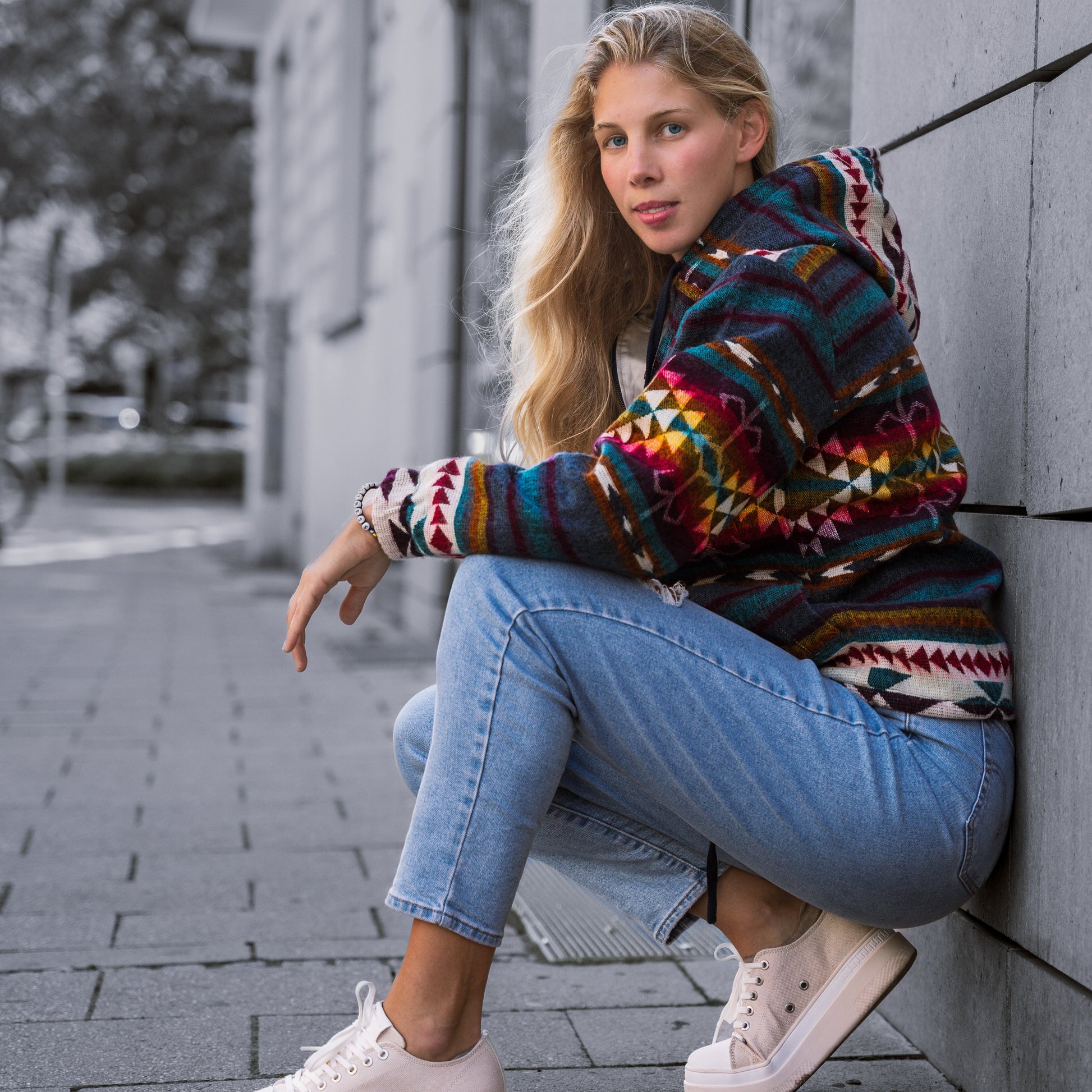 Side view of a woman wearing the QILLU boho jacket made from a wool blend with alpaca, featuring radiant Andean geometric patterns in red, green, yellow, and cream. Photographed outdoors in an urban setting, sitting against a wall.