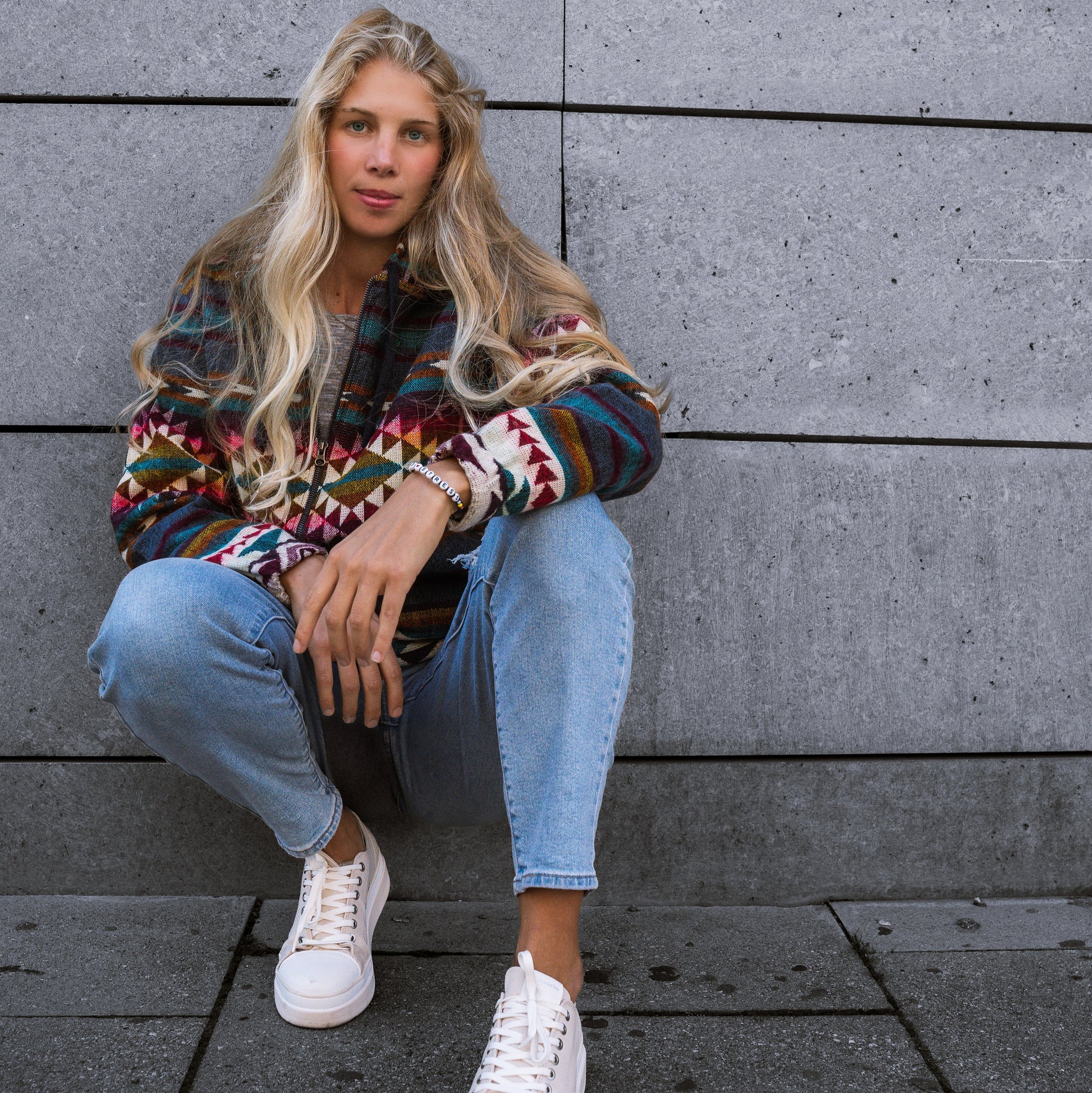 Front view of a woman sitting against a stone wall wearing the QILLU boho jacket made from a wool blend with alpaca, featuring colorful Andean geometric patterns in red, yellow, green, and cream. Photographed outdoors in an urban setting.