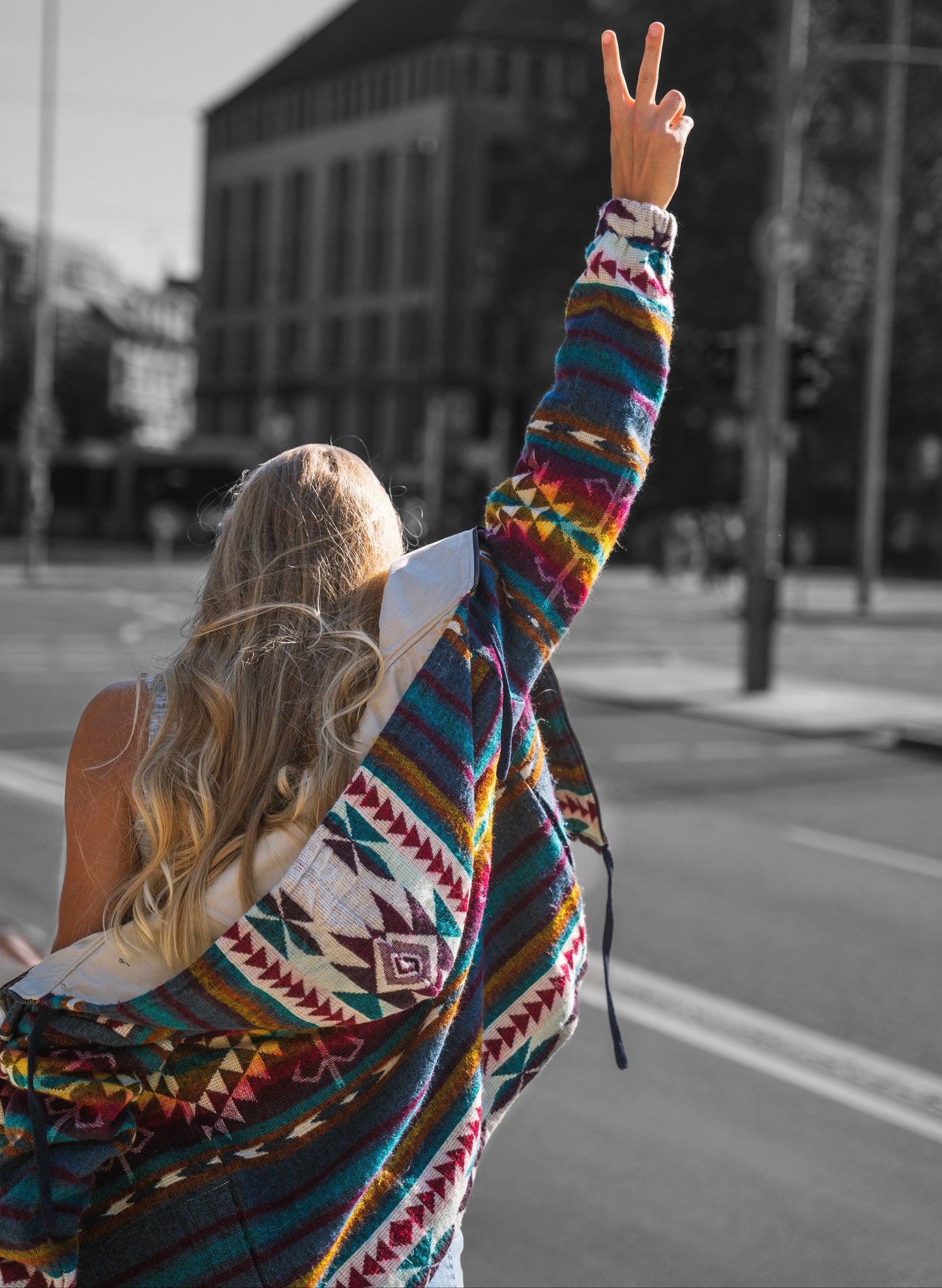Back view of a woman wearing the QILLU boho jacket made from a wool blend with alpaca, showing colorful Andean geometric patterns in red, yellow, green, and cream. She raises her arm making a peace sign while walking in an urban street setting.