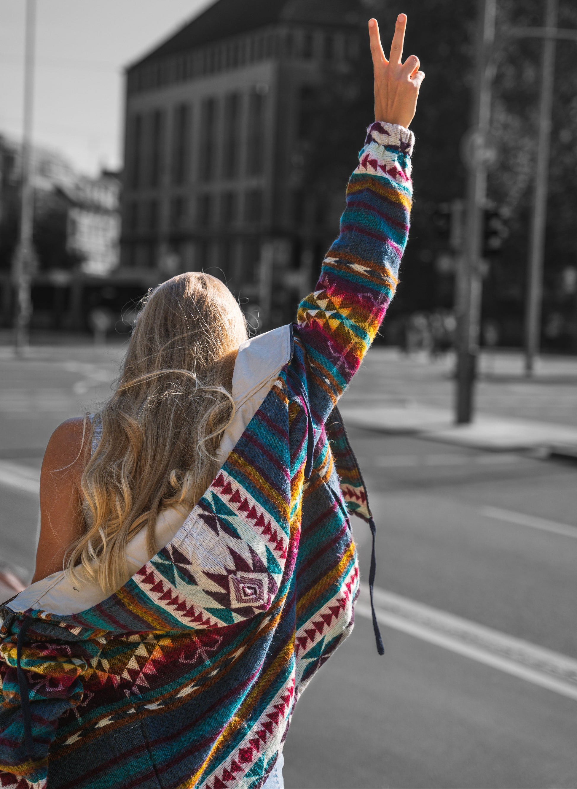 Back view of a woman wearing the QILLU boho jacket made from a wool blend with alpaca, showing colorful Andean geometric patterns in red, yellow, green, and cream. She raises her arm making a peace sign while walking in an urban street setting.