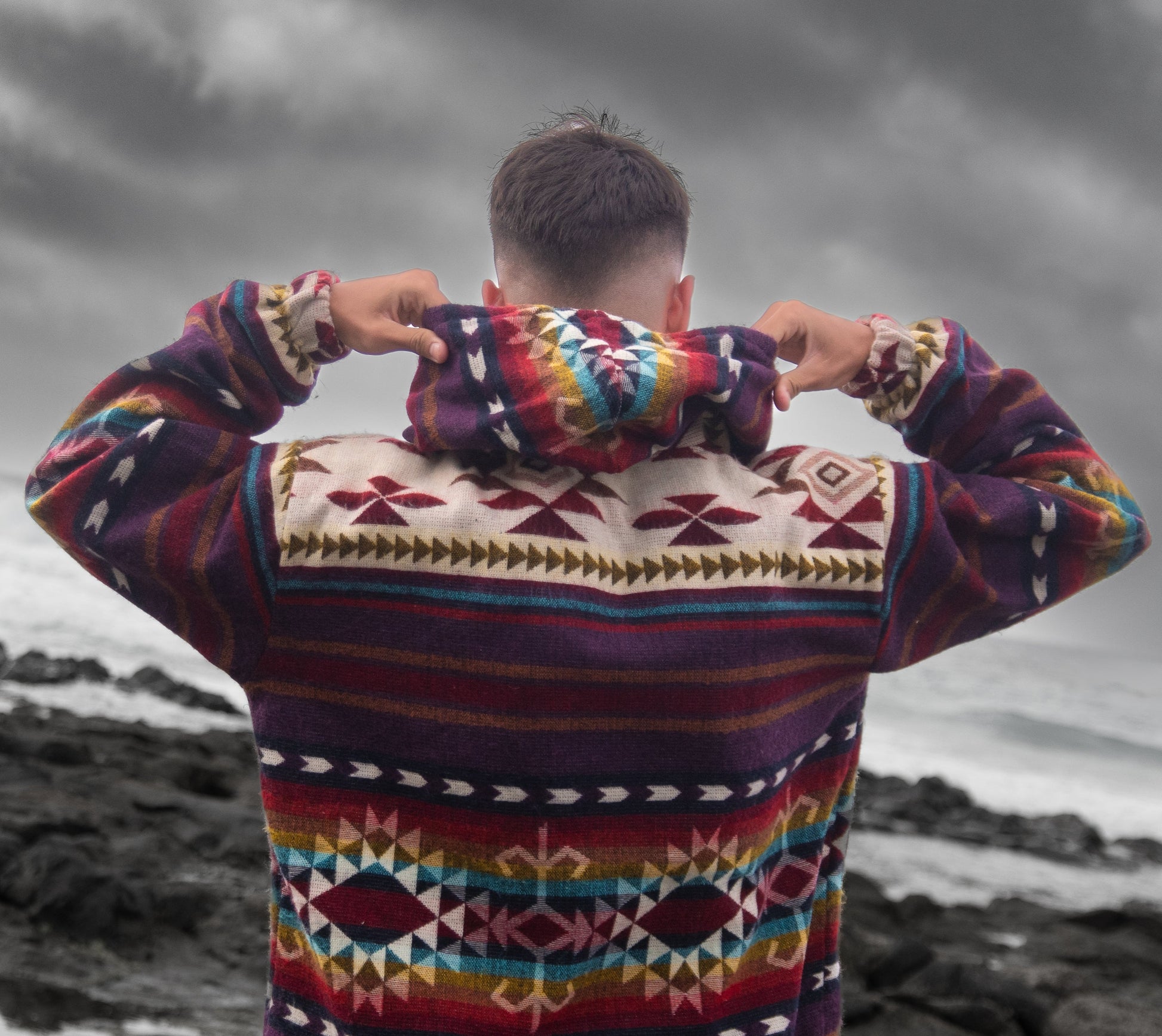 Lifestyle photo of the Chaska jacket by QUENDE – back view of man wearing handmade multicolor wool jacket with hood, ocean background.