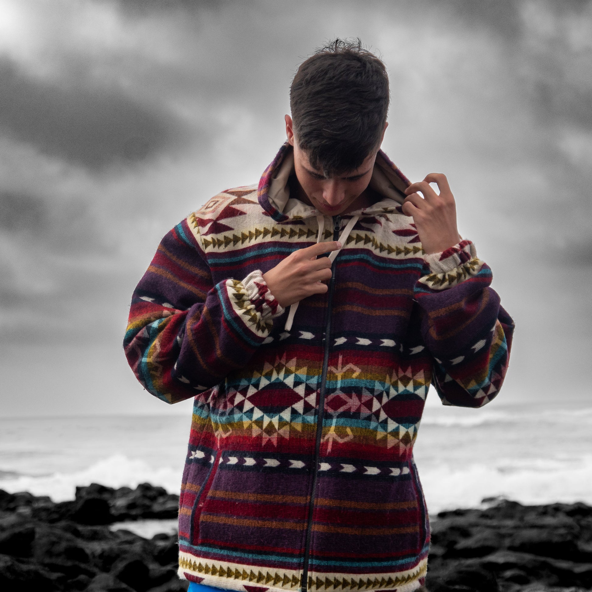 Lifestyle photo of the Chaska jacket by QUENDE – man wearing artisan multicolor wool jacket with hood, ocean background.