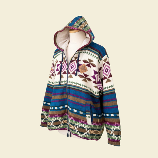 Angled front view of the WAYRA boho jacket made from a wool blend with alpaca, featuring colorful geometric Andean patterns in blue, green, purple, brown, and cream, with a hood and zipper. Photographed against a solid cream background.