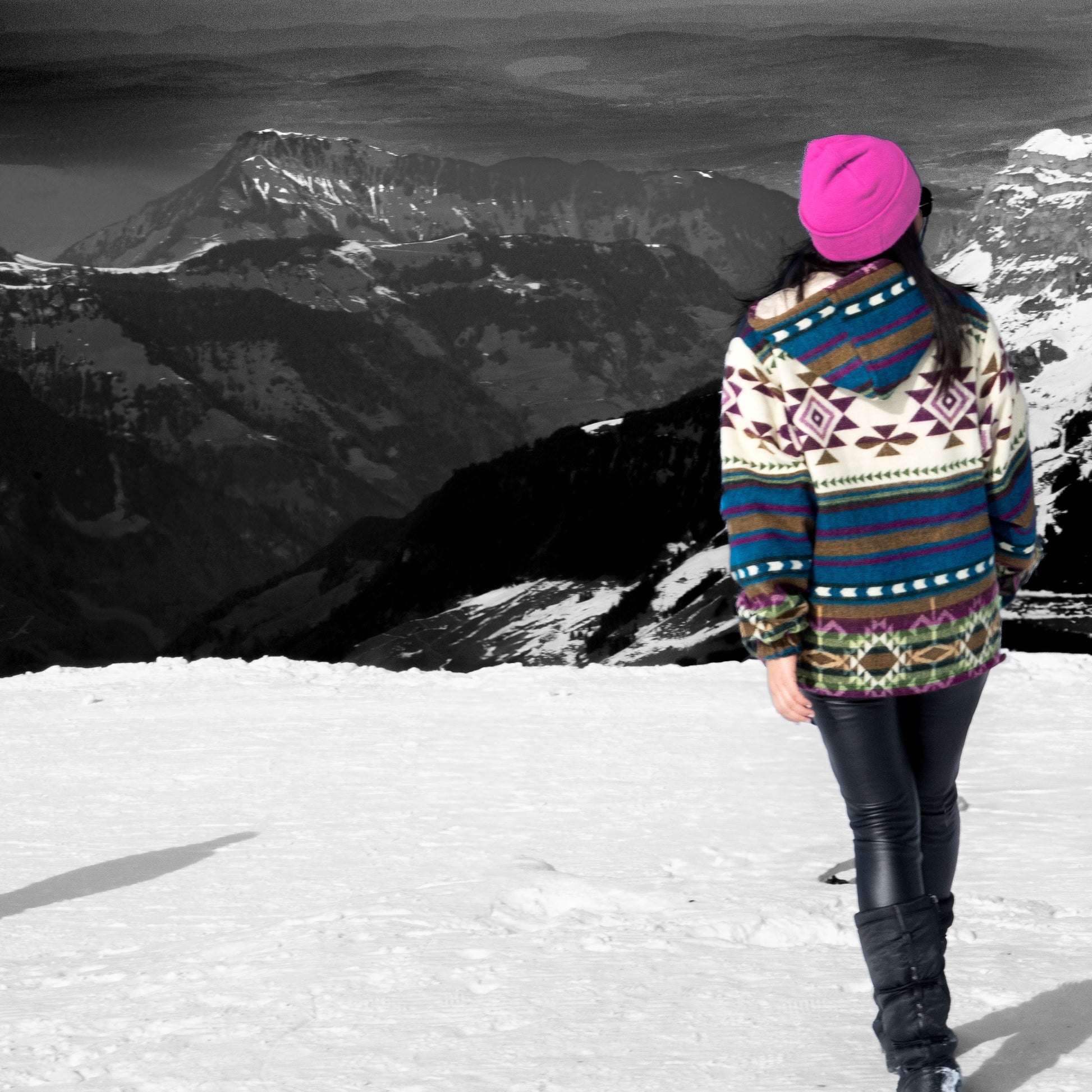Back view of a woman wearing the WAYRA boho jacket made from a wool blend with alpaca, featuring geometric Andean patterns in blue, green, purple, brown, and cream. Photographed outdoors in the mountains, with snow and a pink beanie adding contrast.